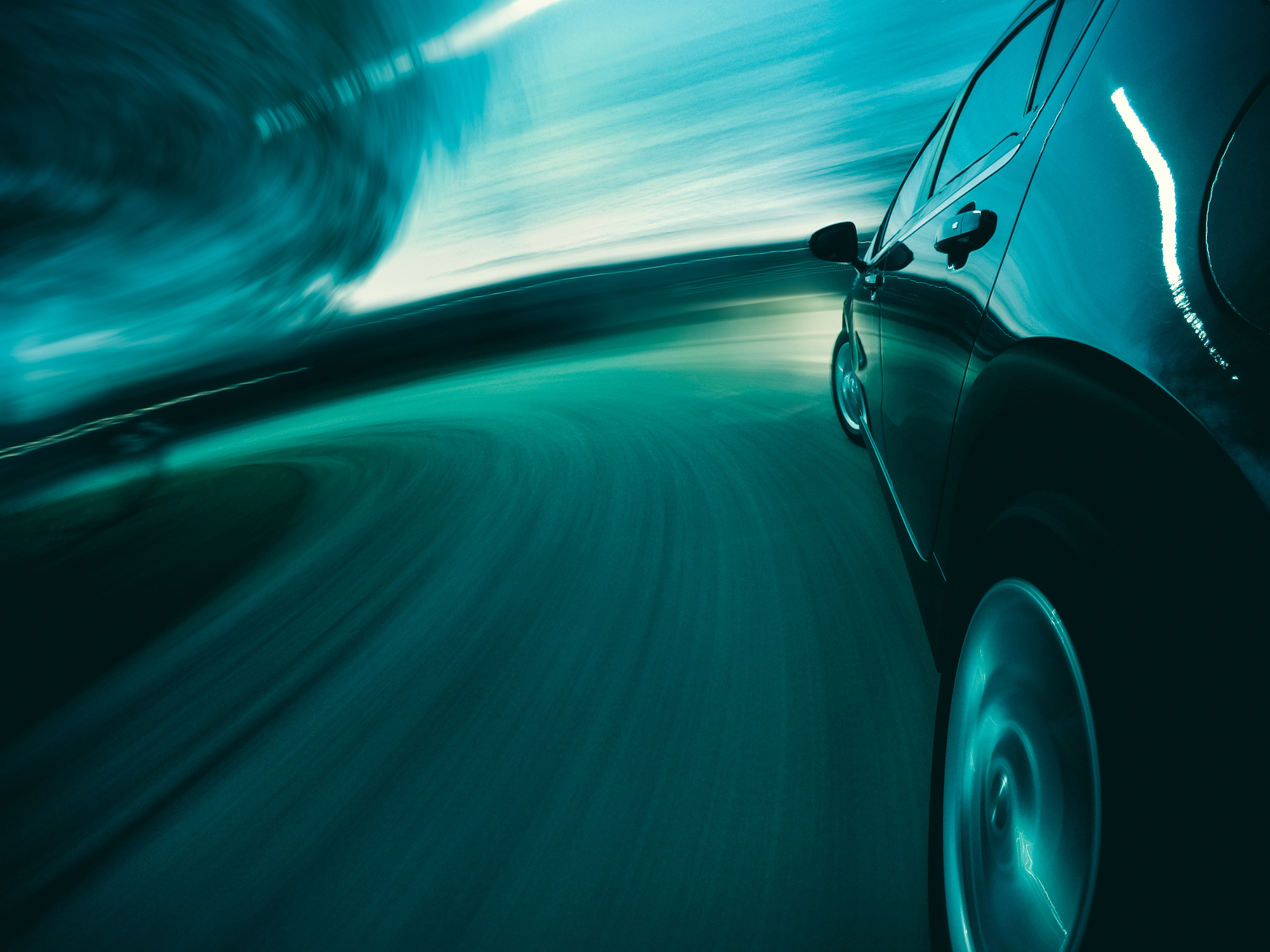 A dark car speeds through a teal and turquoise blurred tunnel, captured from a low, dynamic angle showing motion blur on the wheels and background.